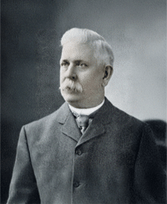 An older man with white hair and a large white mustache, wearing a dark suit and high-collared shirt, poses for a formal portrait against a plain background.