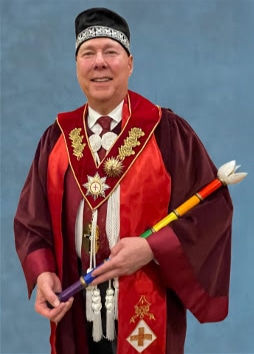 A smiling man in ceremonial maroon and red robes with gold decorations, wearing a black hat, and holding a colorful staff, standing against a plain blue background.