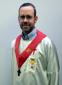A man wearing glasses and a white religious robe with a red stole, a black cross necklace, and gold embroidered symbols stands against a plain light background.